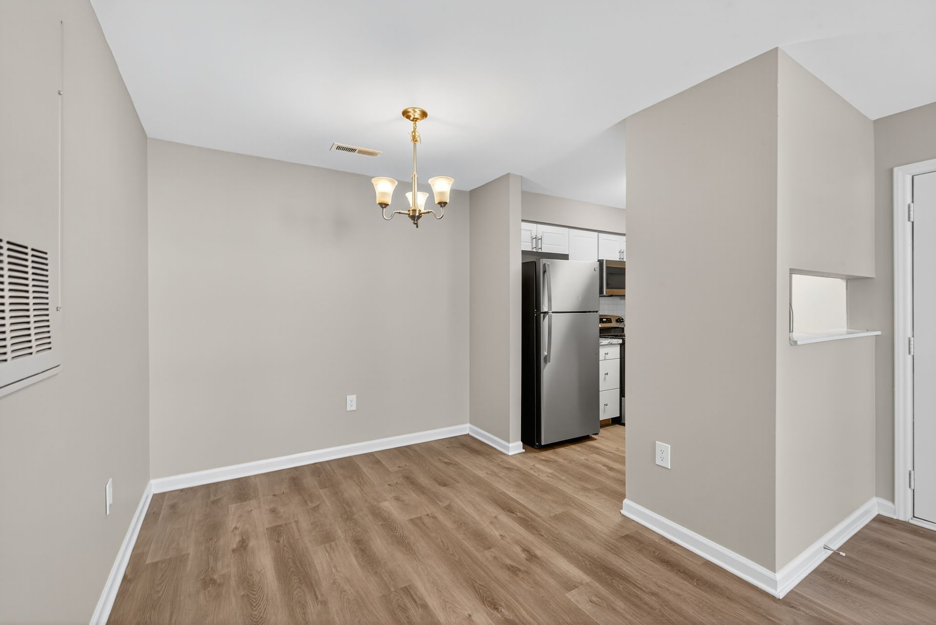 Empty dining area with light gray walls, wood-look flooring, and a chandelier near a kitchen opening