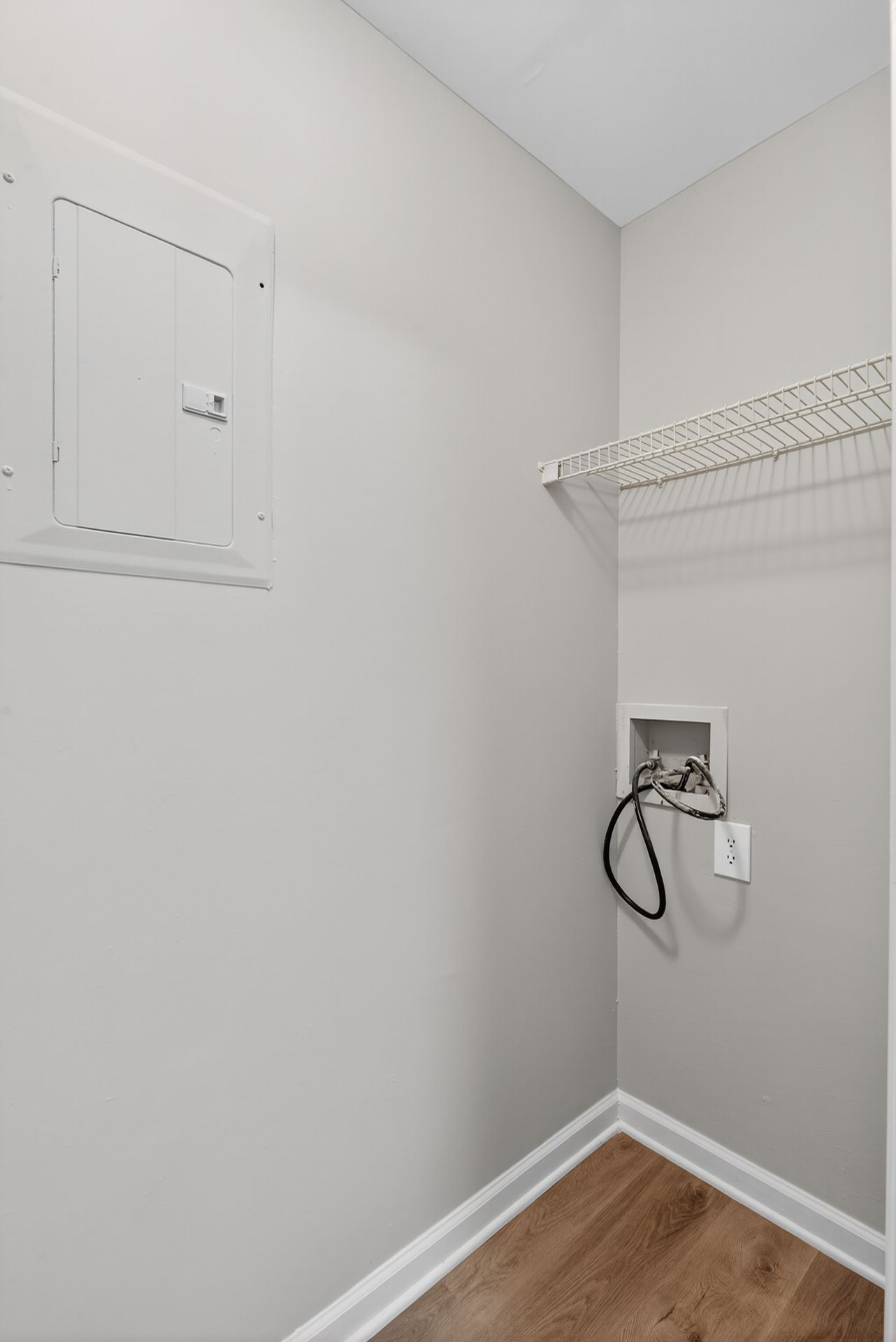 Small laundry/utility closet with white walls, wire shelf, electrical panel, hookups, and wood floor.