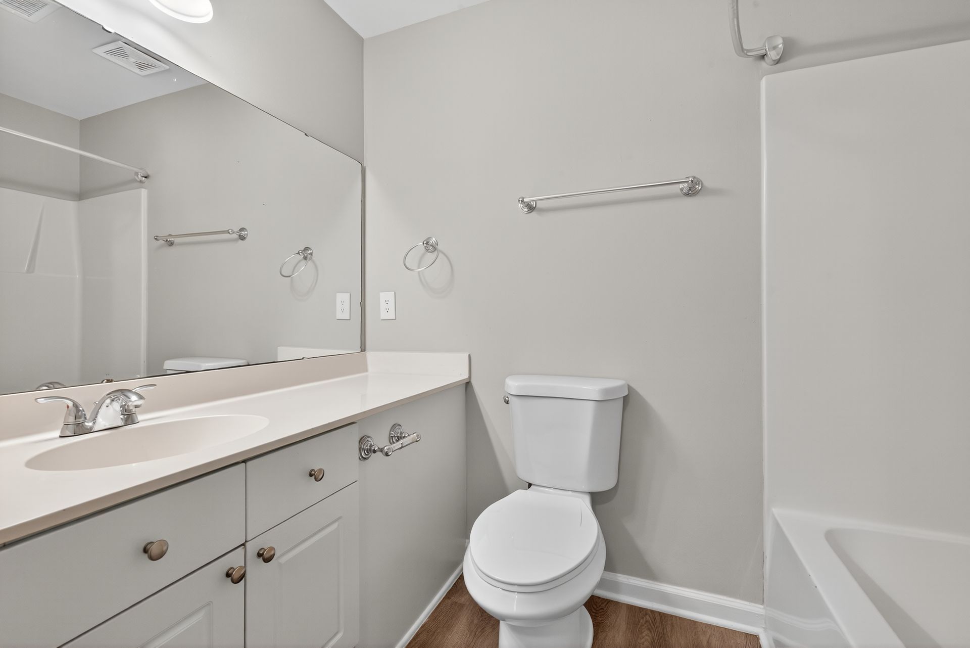 Bright bathroom with white vanity, toilet, mirror, and tub against light gray walls.