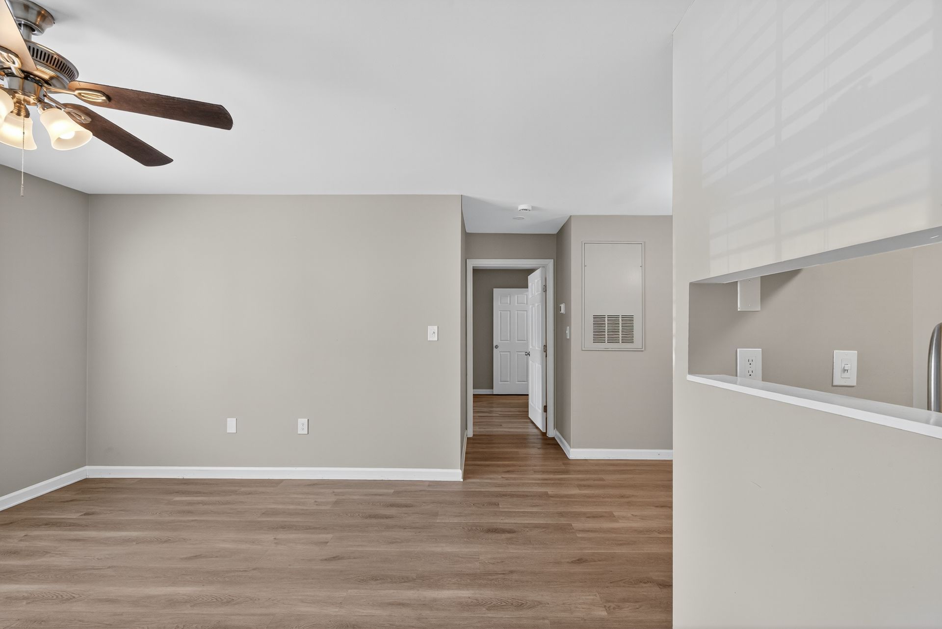 Empty living room with gray walls, wood floors, ceiling fan, and a pass-through opening to the kitchen.