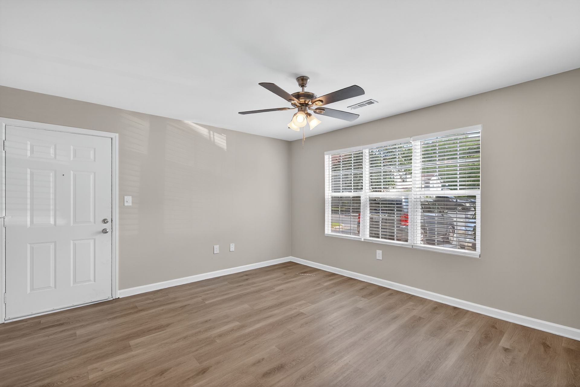Empty bedroom with gray walls, wood flooring, white closet doors, and a window with blinds and ceiling fan.