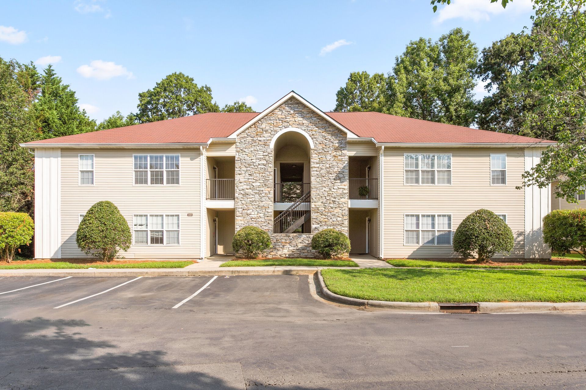 Two-story beige apartment building with red roof and central stone entrance, viewed from the parking lot