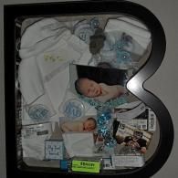 Shadow-box display with baby clothes, photos, and keepsakes arranged in a black frame.