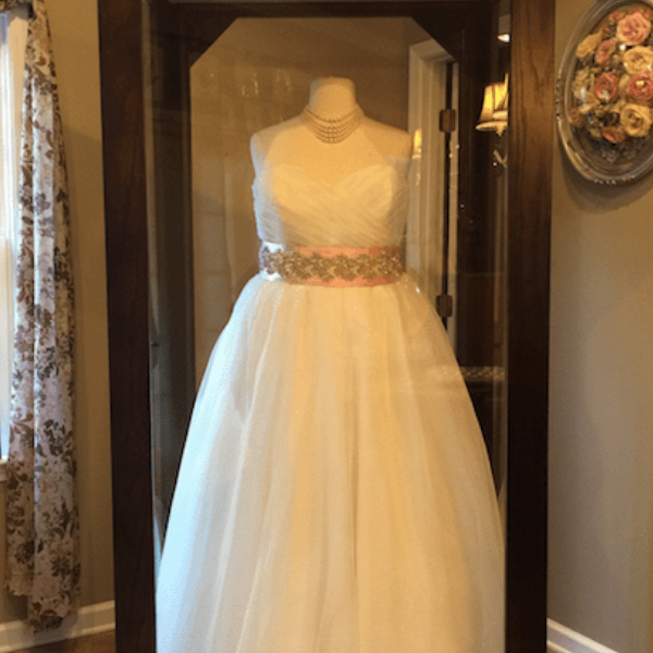 White sleeveless bridal gown on a mannequin in a display case, with a floral waist belt