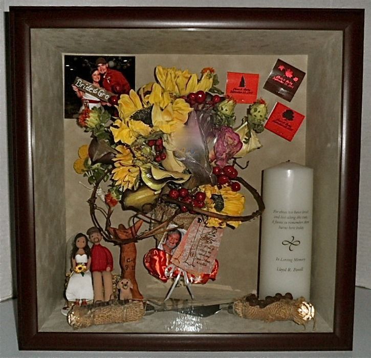 Framed floral memorial display with yellow and red flowers, figurines, a candle, and small keepsakes