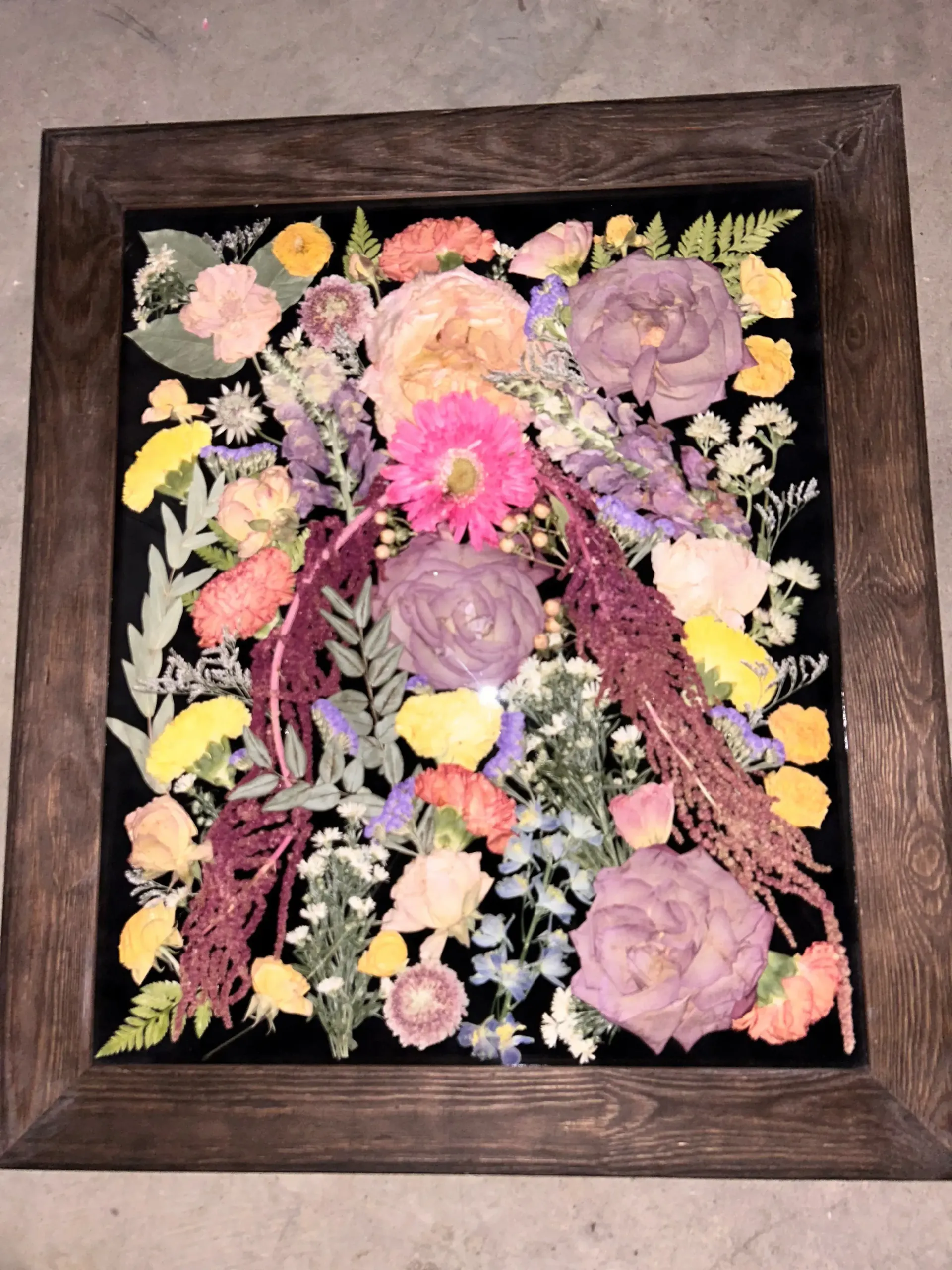 Pressed flower art in a dark wooden frame with pink, purple, and yellow blooms on black background