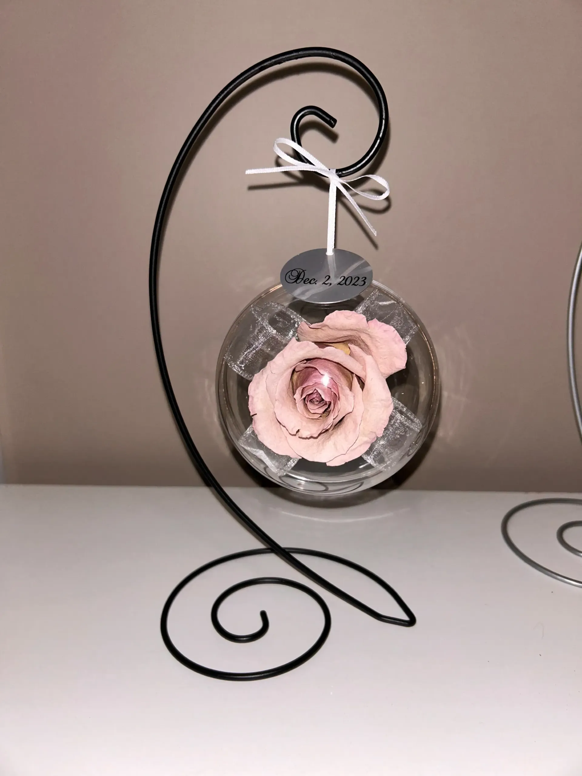 Pink rose in a round glass ornament hanging from a black metal stand on a table.