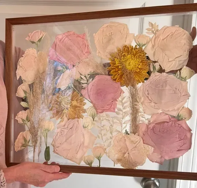 Framed pressed flowers in soft pinks and beige with a yellow bloom, held in two hands