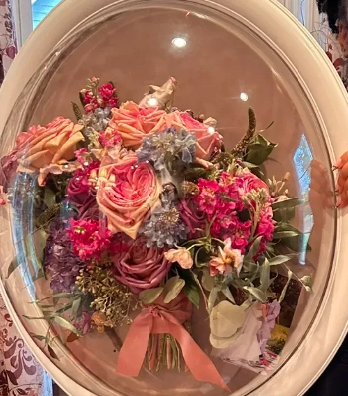Pink and purple floral bouquet wrapped in clear cellophane with a peach ribbon, displayed in an oval frame