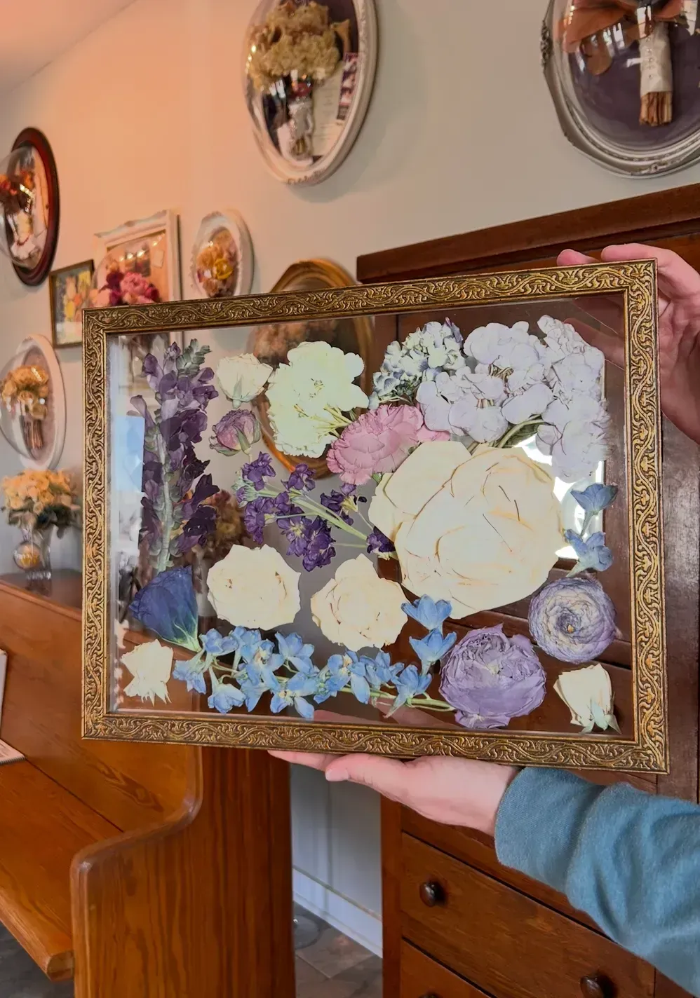 Framed pressed flower arrangement held in a hand, with pastel blooms and purple accents.