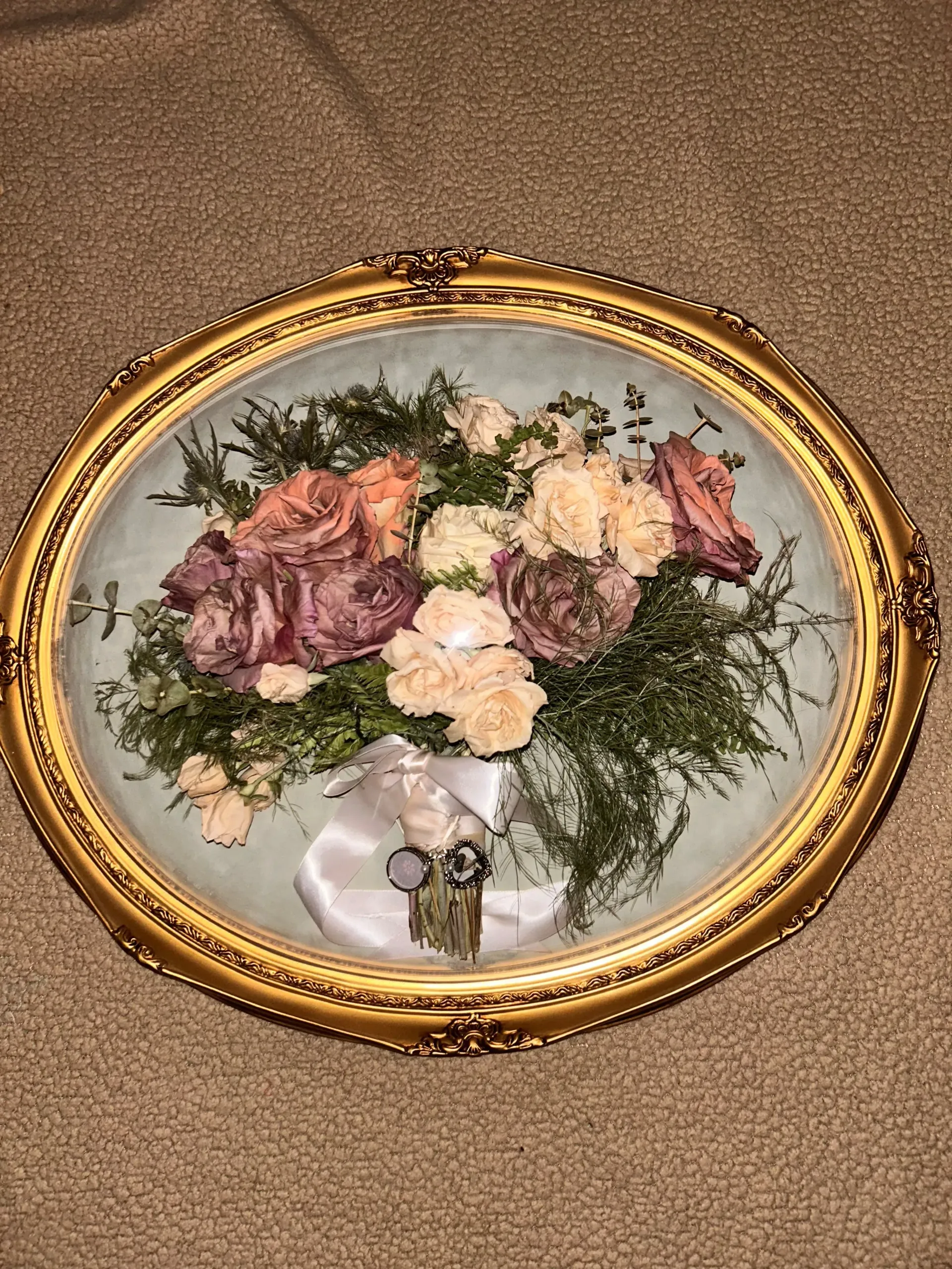 Oval gold-framed floral arrangement with pink and cream flowers on a pale background, resting on carpet