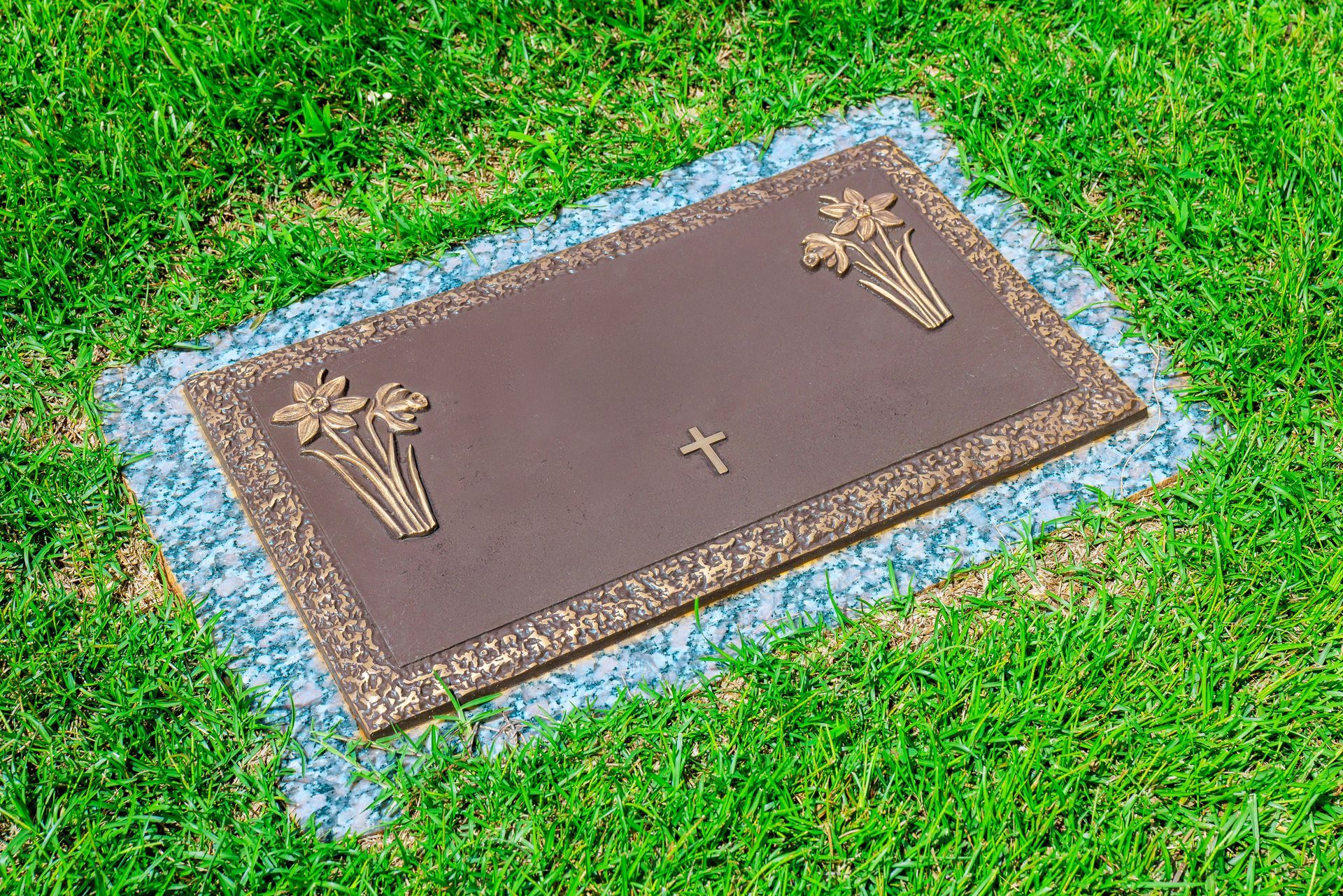 A flat bronze grave marker with floral relief and cross, mounted on a granite base