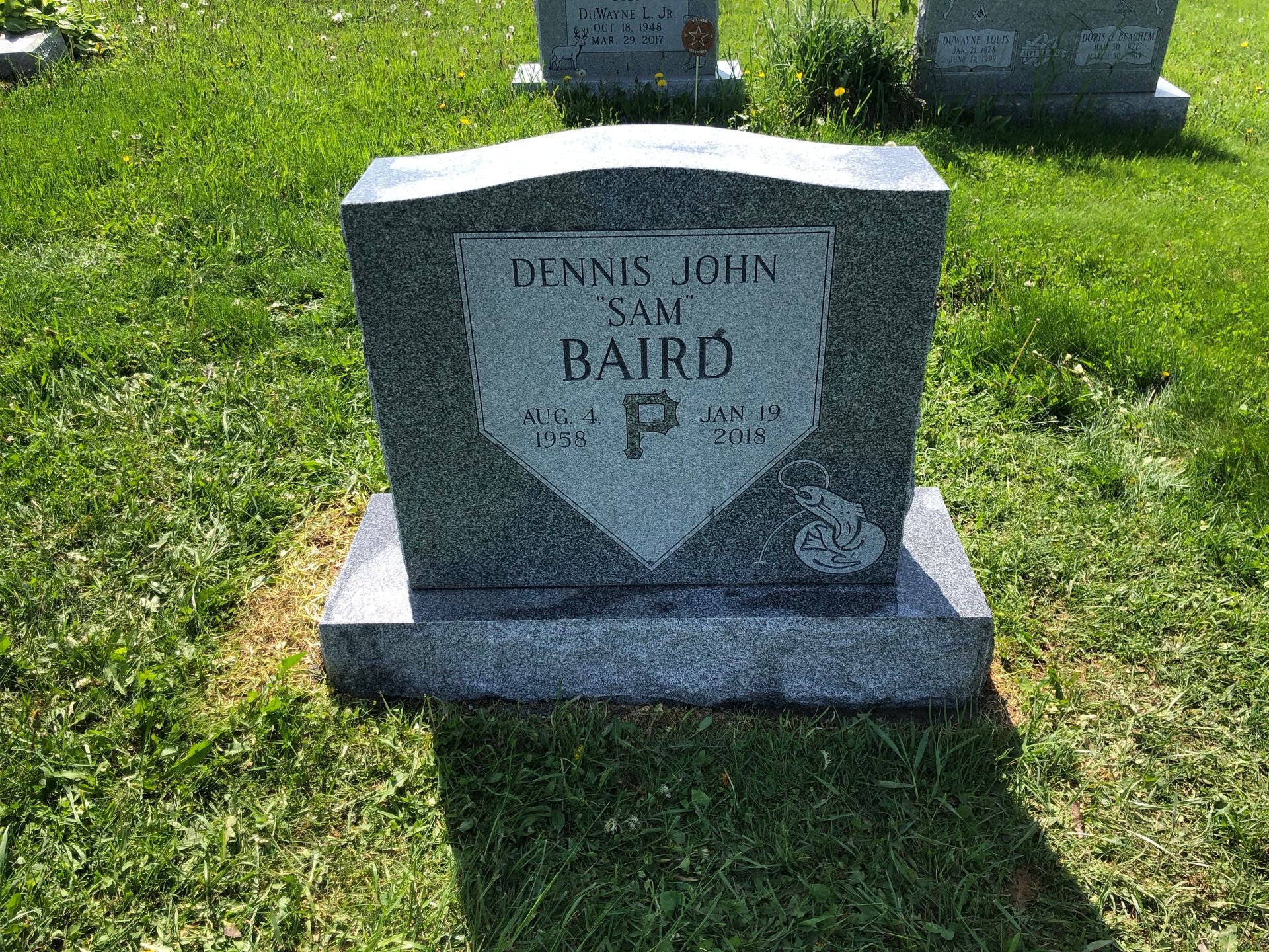 Gray granite gravestone in a sunny green cemetery, engraved with the name Dennis John Baird.