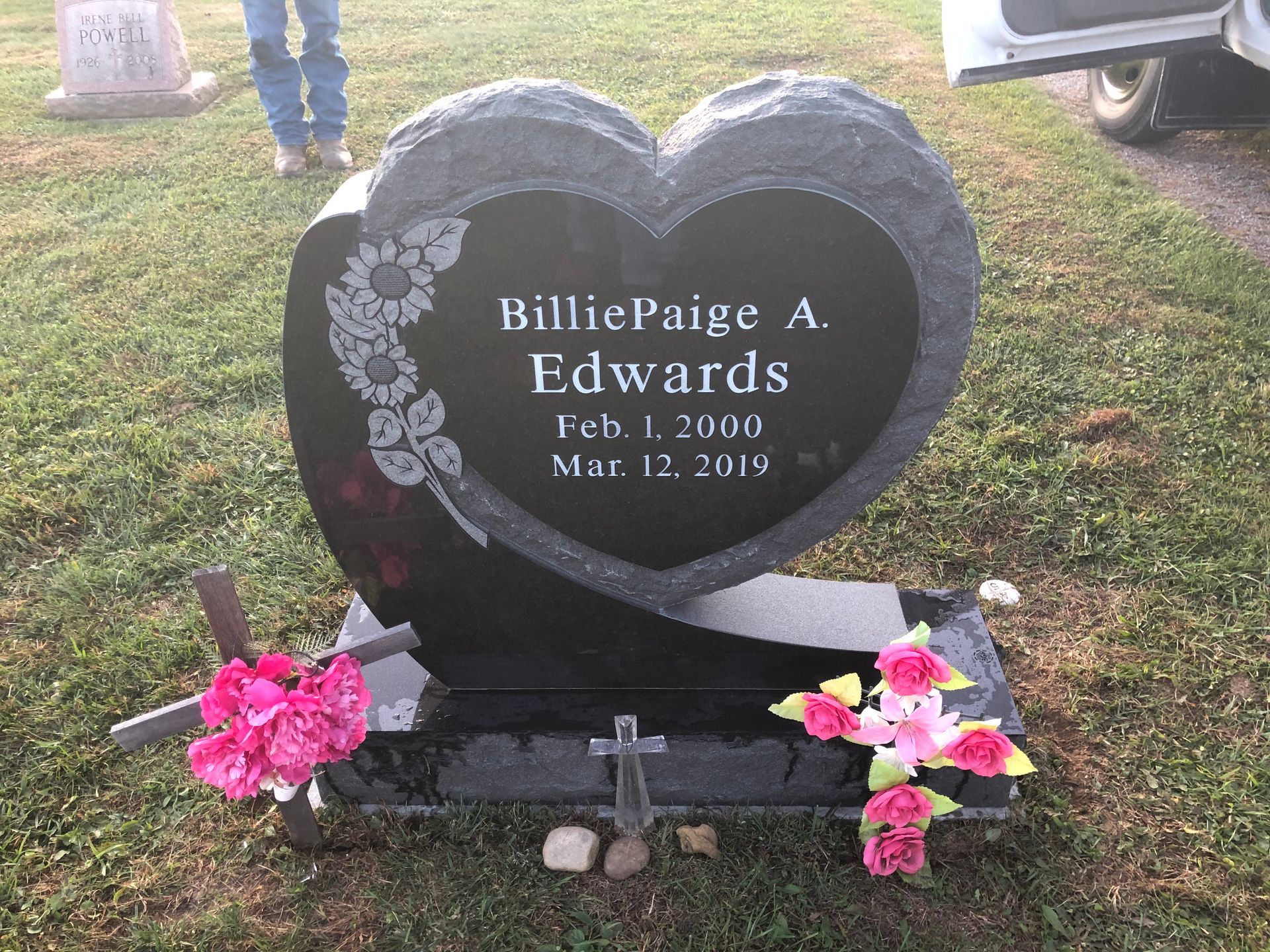 Heart-shaped black gravestone with pink flowers and a cross in a grassy cemetery.