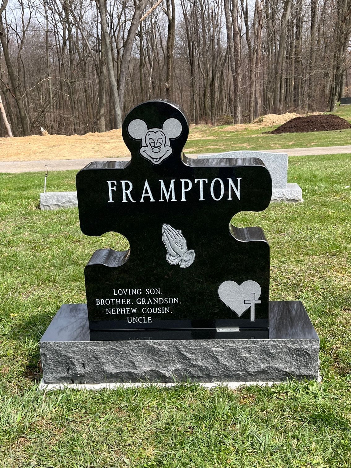 Black gravestone shaped like a puzzle piece reading “FRAMPTON” in a grassy cemetery.