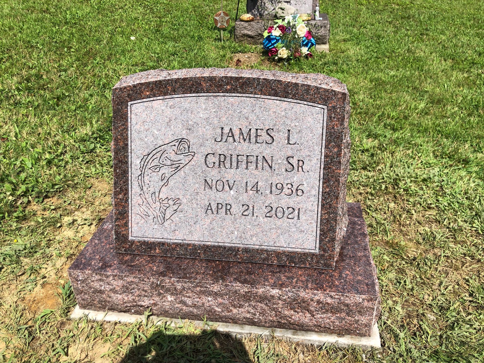 Gray granite gravestone in grass with engraved name, dates, and small flowers above it