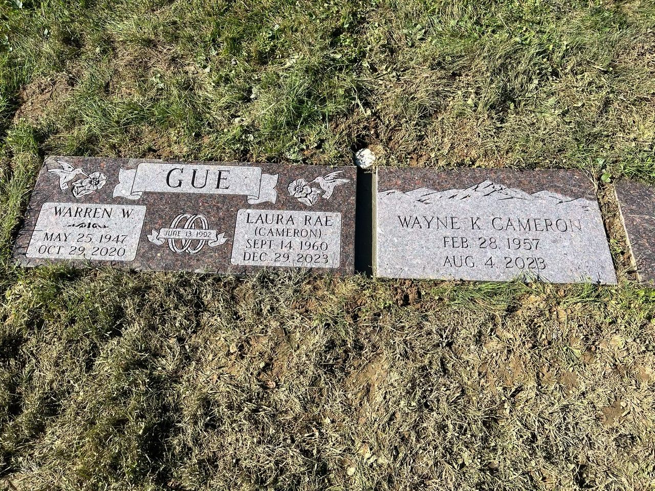 Three weathered grave markers on grass, including one for “GUF” and two small plaques.