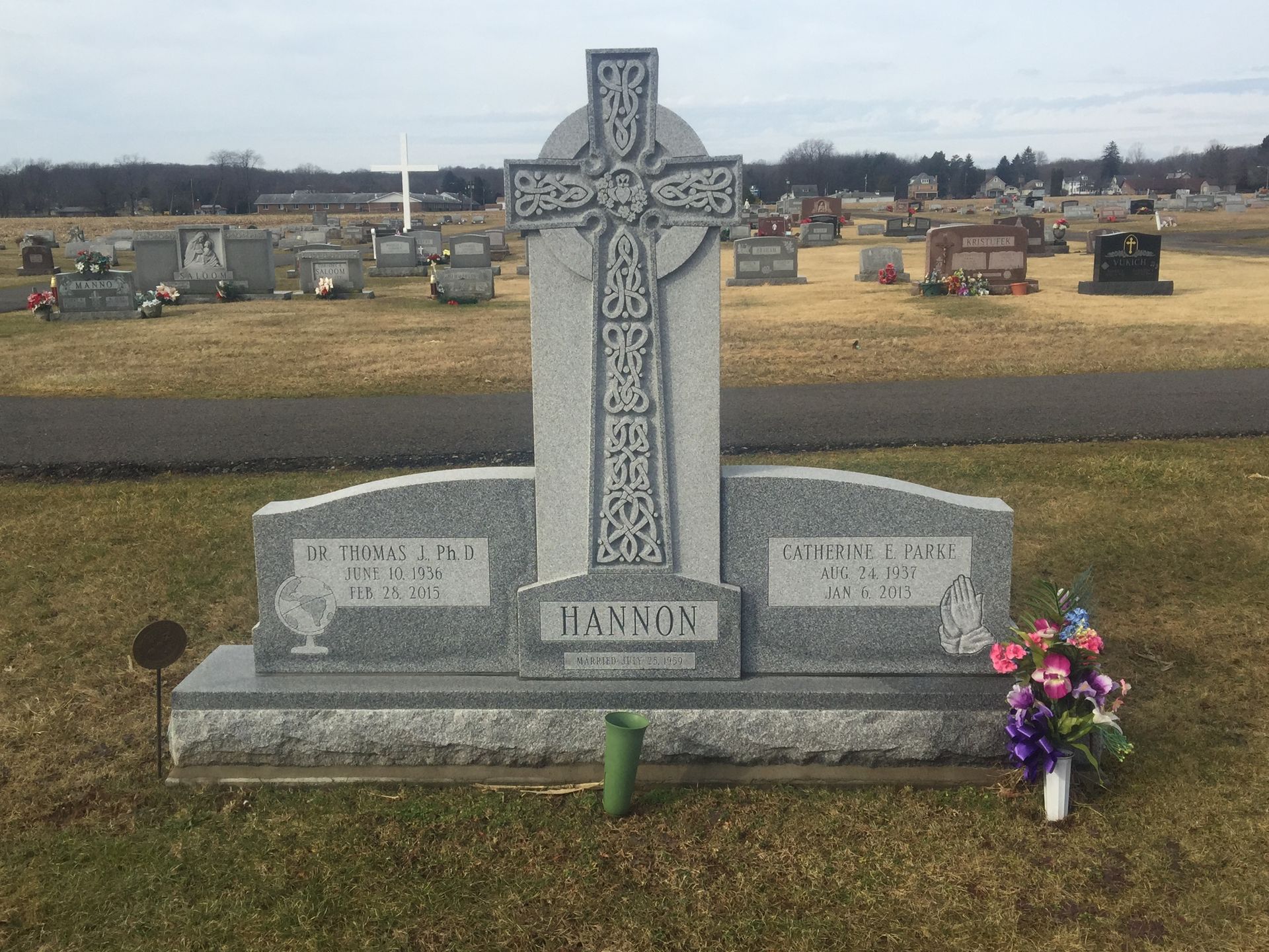 Gray memorial headstone with a cross in a cemetery, with flowers at the base.