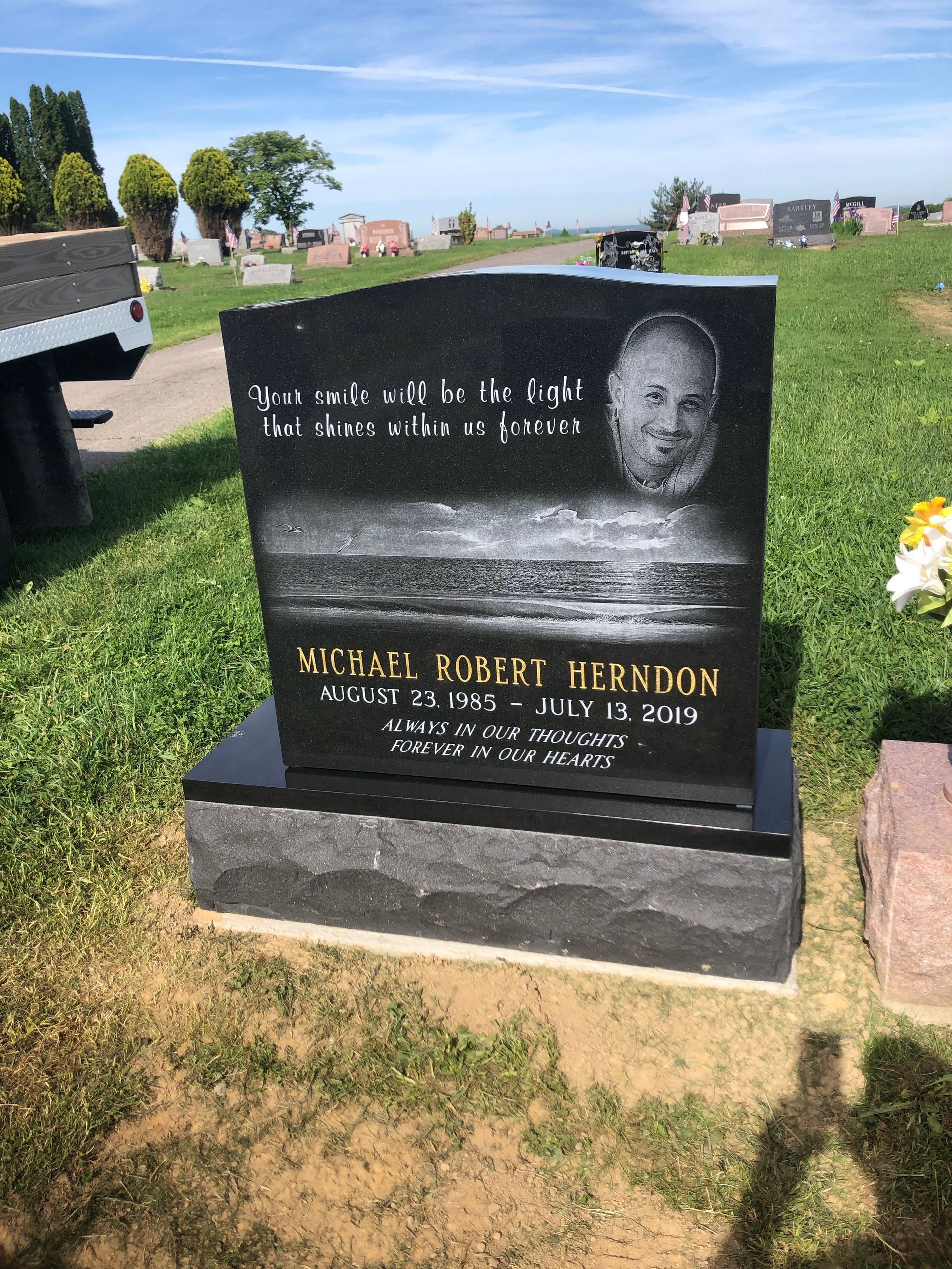Black granite memorial headstone in a grassy cemetery, with engraved portrait and gold lettering