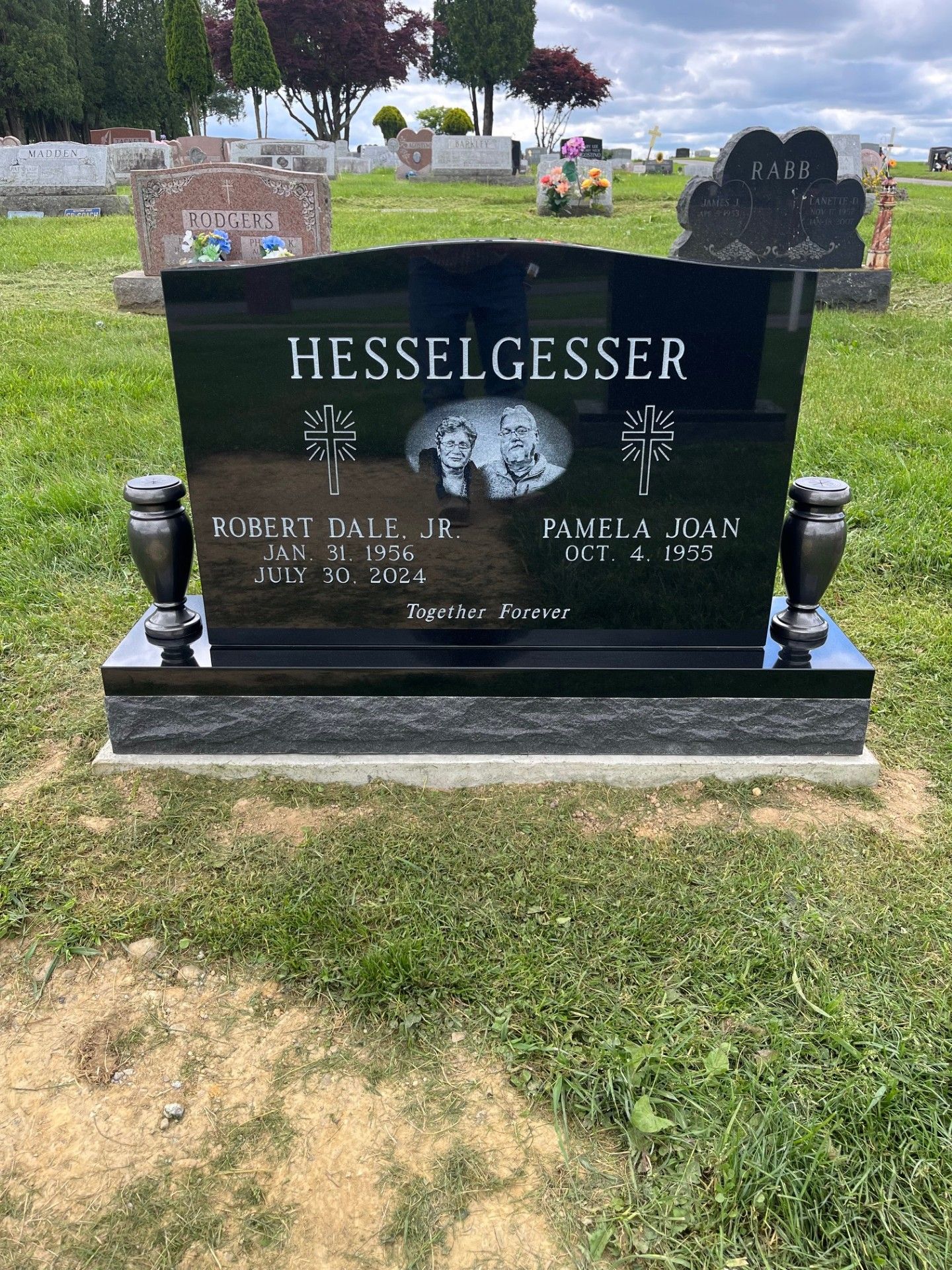 Black granite headstone in a cemetery with a portrait and engraved names, surrounded by grass and nearby graves
