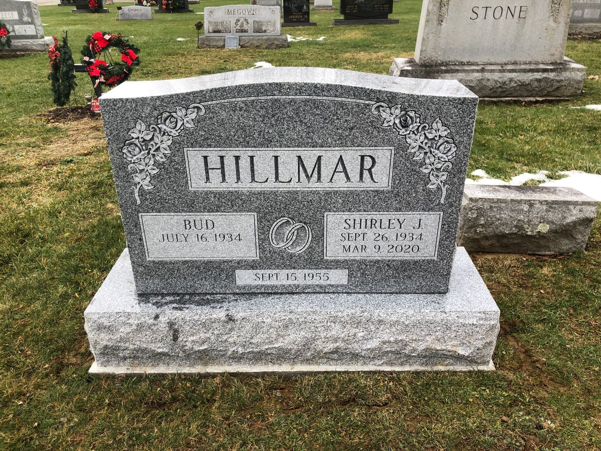 Gray gravestone engraved “HILLMAR” in a cemetery with flowers in the background