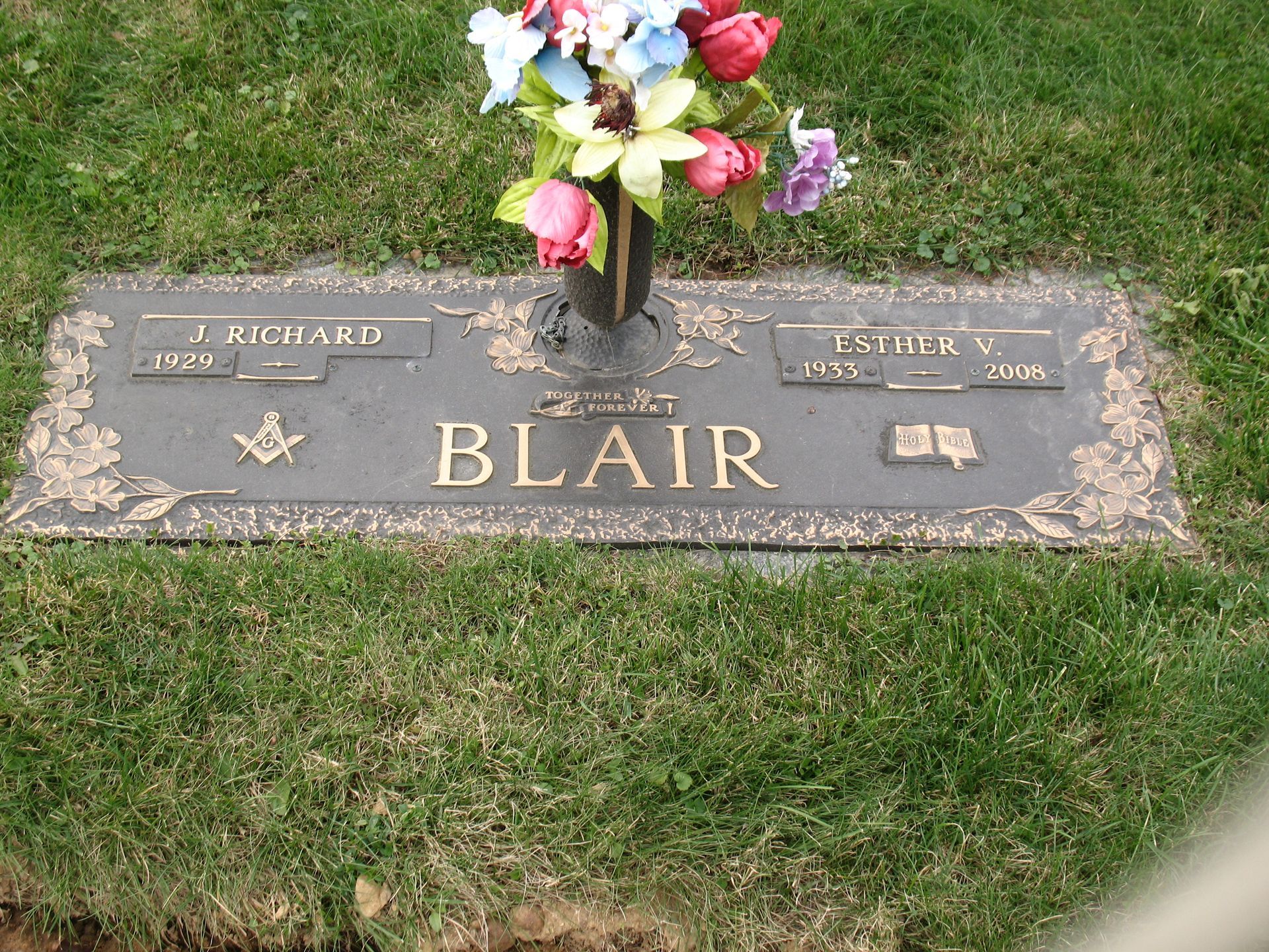Floral arrangement above a gray grave marker engraved “BLAIR” in a grassy cemetery plot