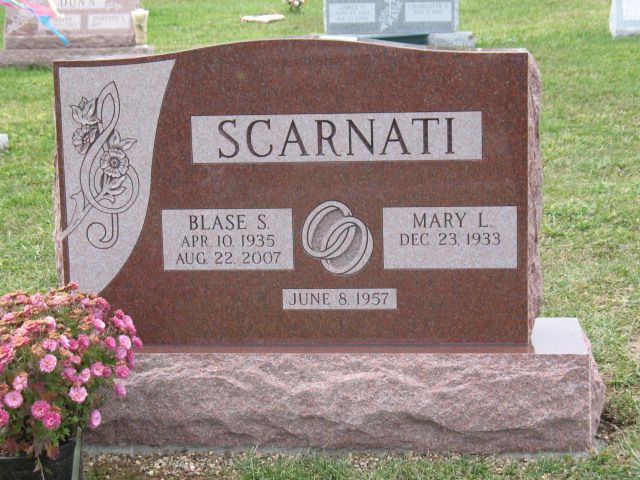 Brown gravestone with “Scarnati” engraved, in a cemetery with pink flowers beside it.