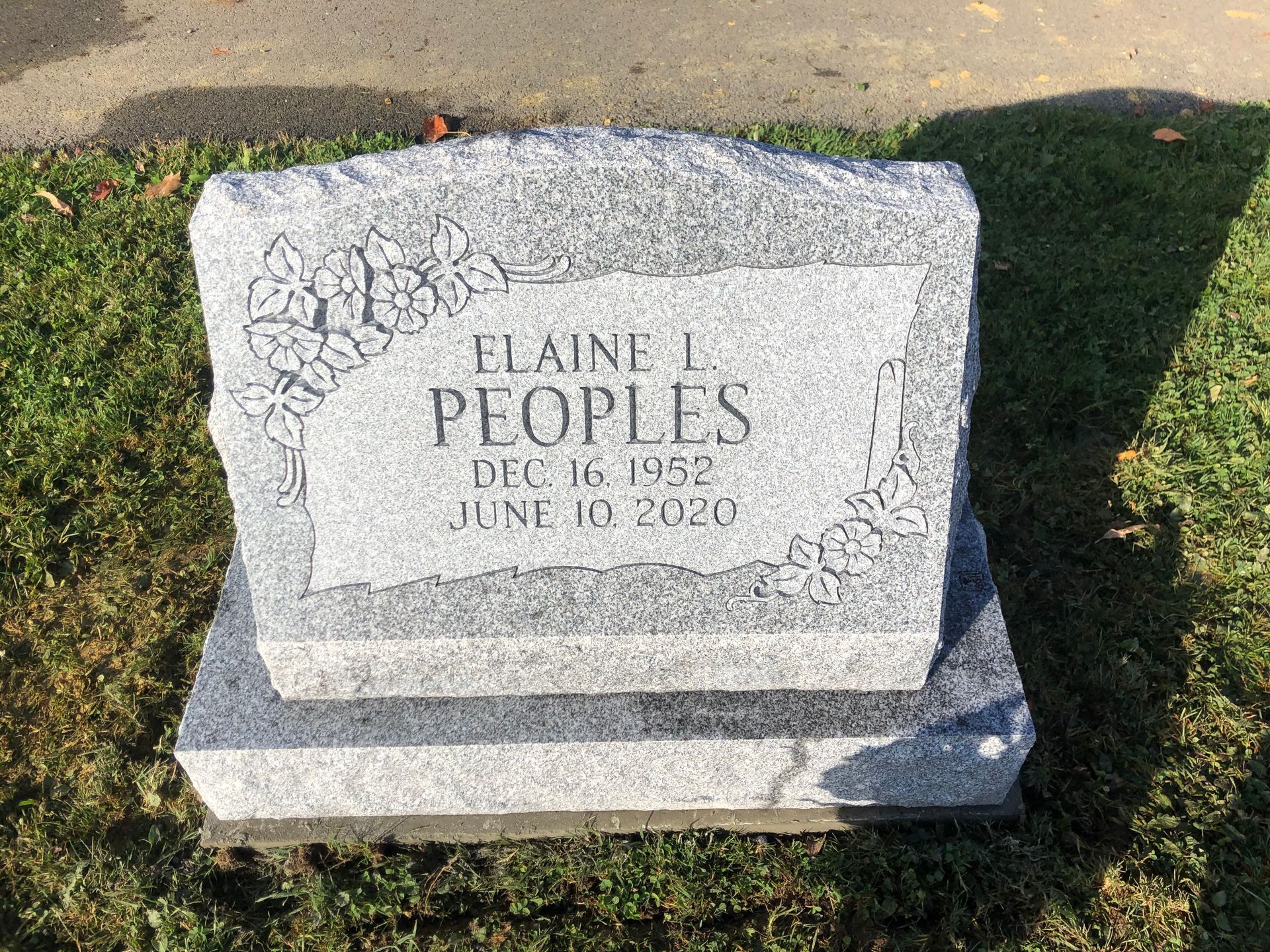 Gray granite gravestone in grass, engraved with “Elaine L Peoples” and a floral design.