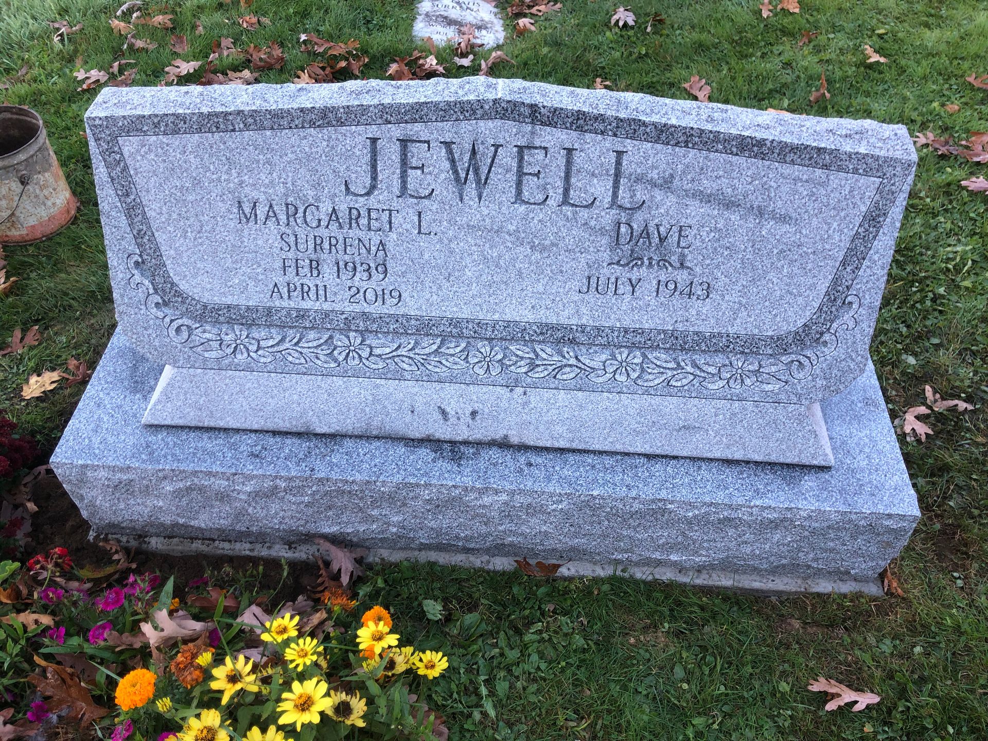 Gray memorial headstone in a flowered cemetery plot with engraved name and dates.