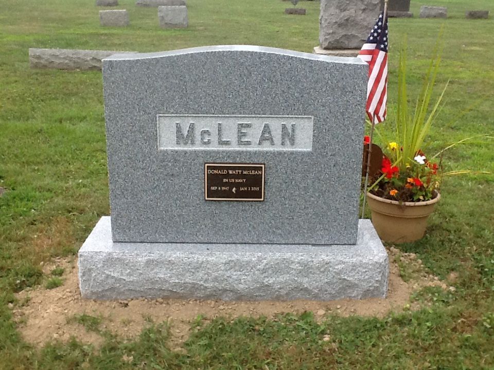 Gray McLean gravestone with a small plaque, U.S. flag, and flower arrangement in a cemetery