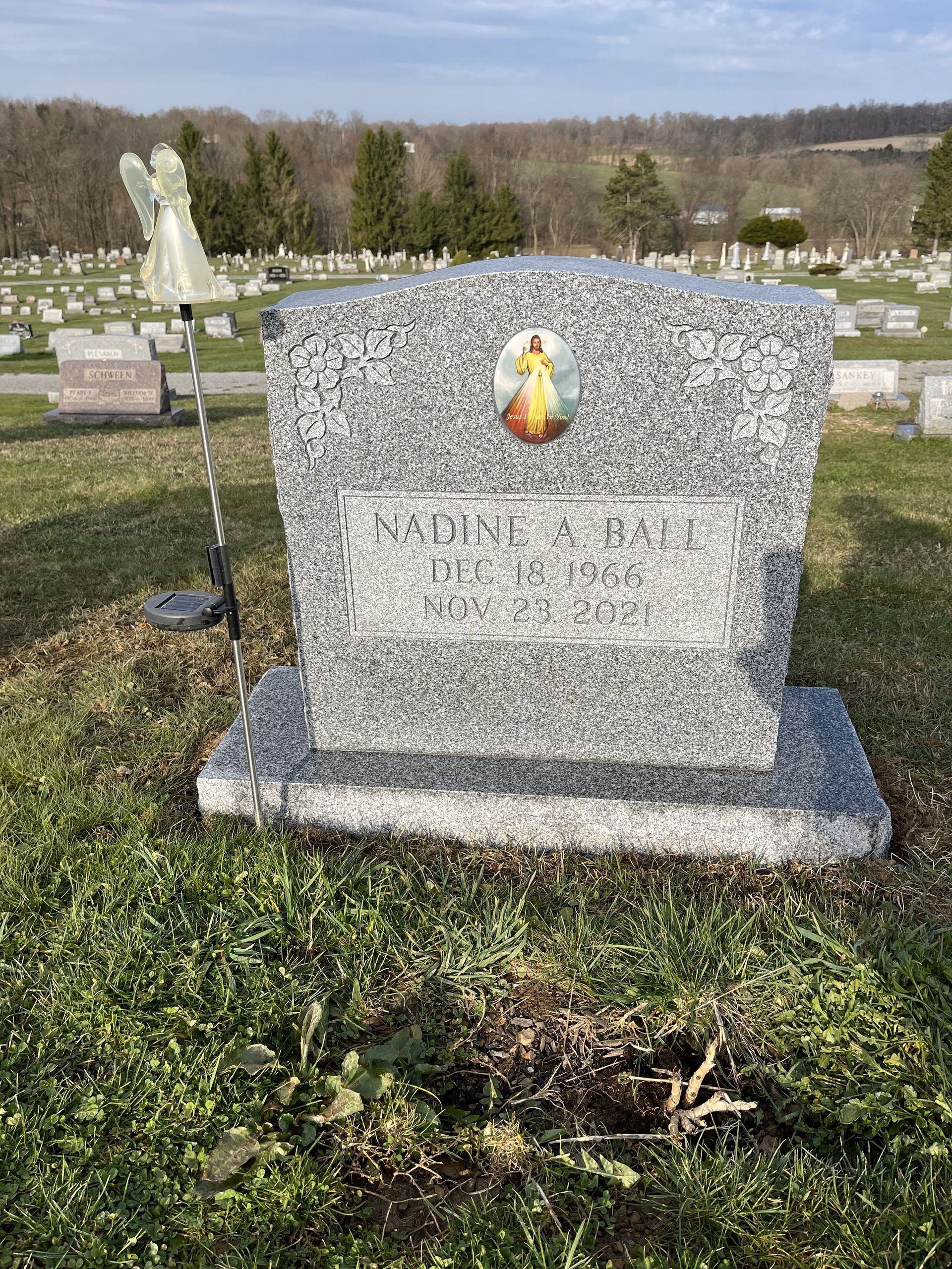 Gray granite gravestone with a small saint plaque in a grassy cemetery on a sunny day