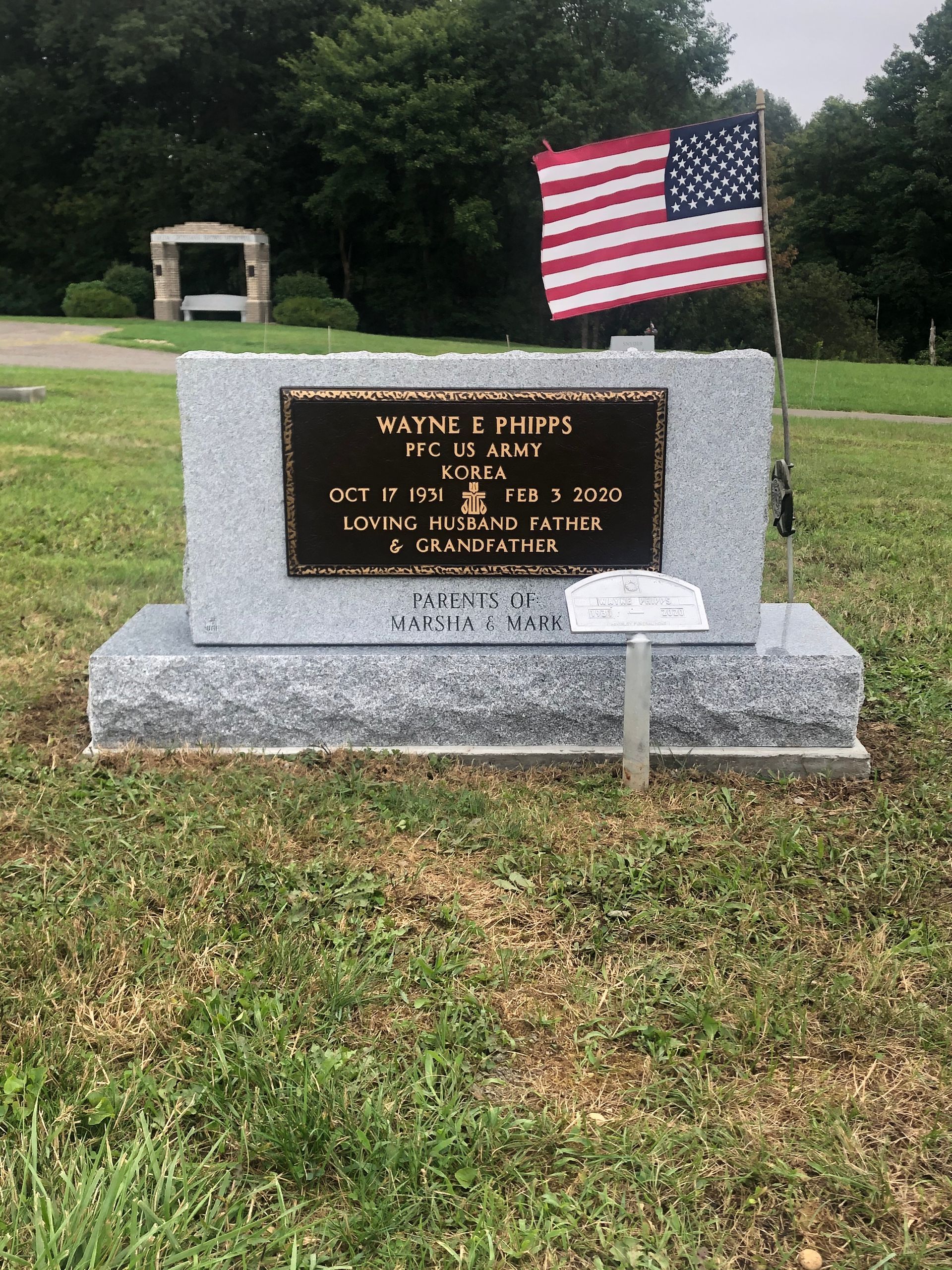 Memorial gravestone with an American flag in a grassy cemetery, with a small shelter in the background