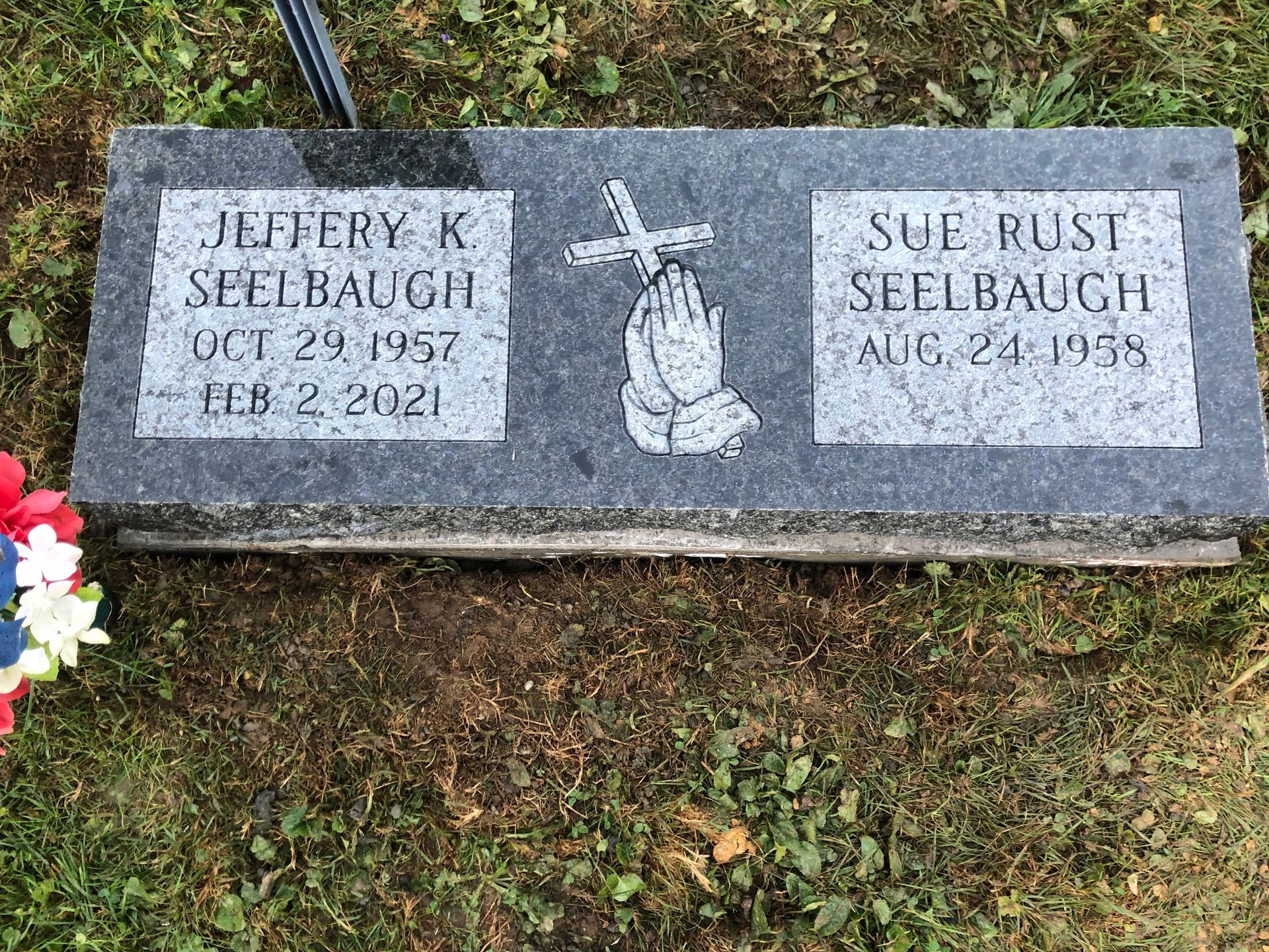 Gravestone with two engraved name plaques, flowers at left, and grass below.