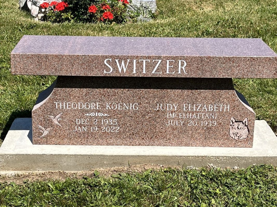 Granite bench in a cemetery, other headstones and flowers visible in the background.
