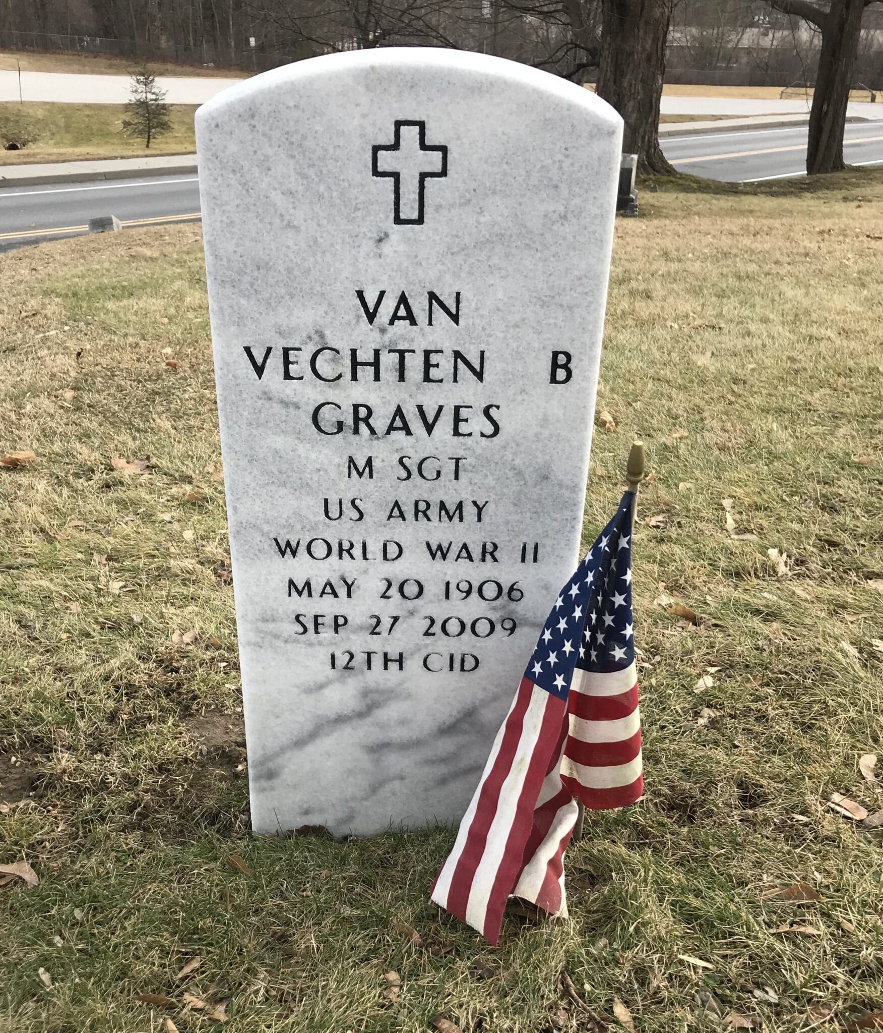 Gravestone with cross, U.S. Army inscription, and small American flag in a cemetery grass yard.