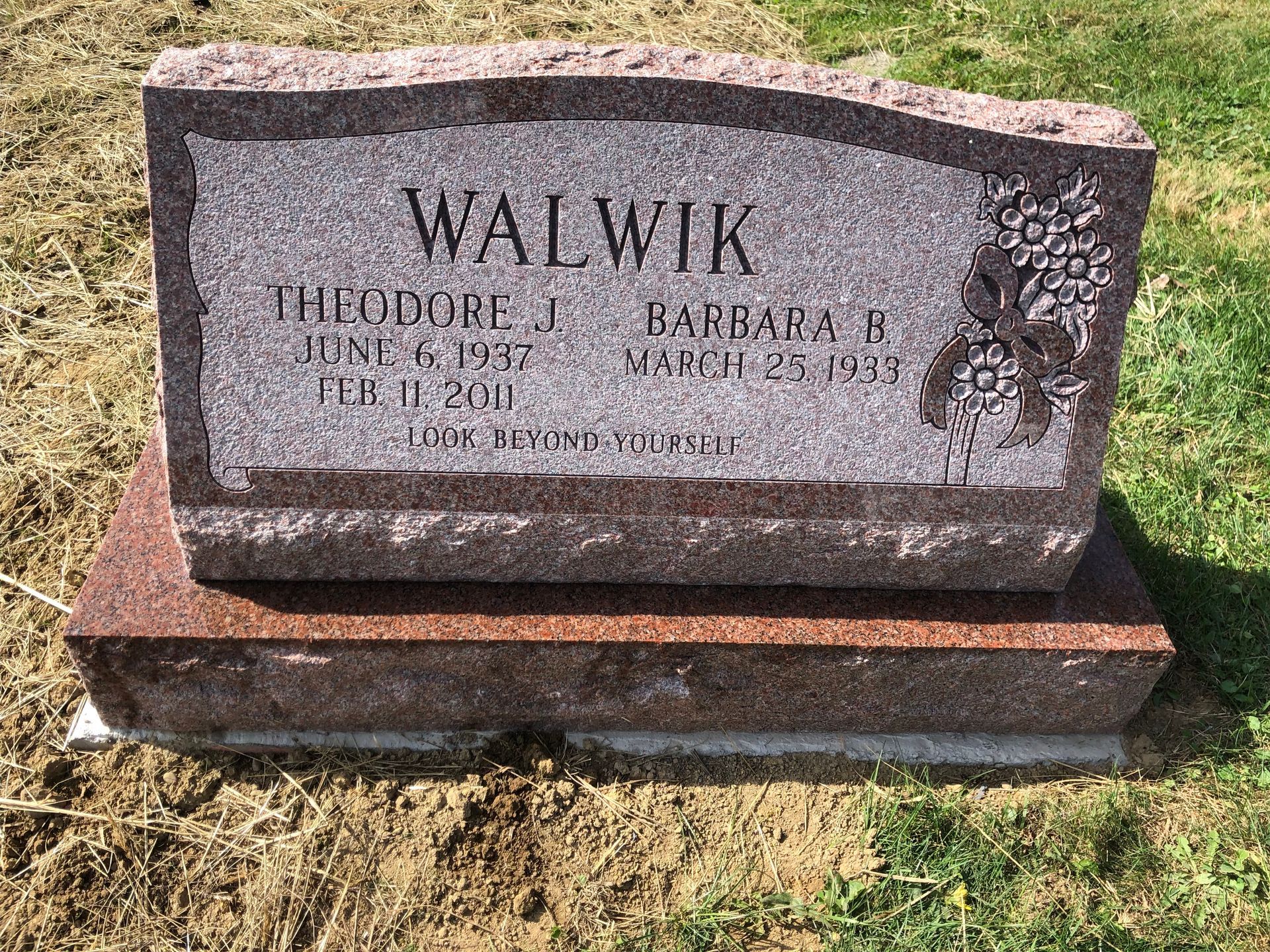 Gray granite gravestone engraved “WALWIK,” on a red base in a grassy cemetery.