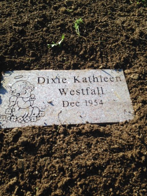 Gravestone plaque reading “Dixie Kathleen Westfall, Dec 1954” in soil with a small angel engraving