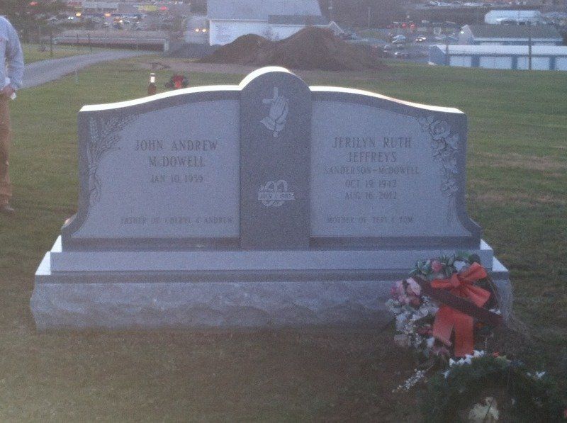 Two-sided memorial gravestone in a grassy cemetery with red flowers at the base.