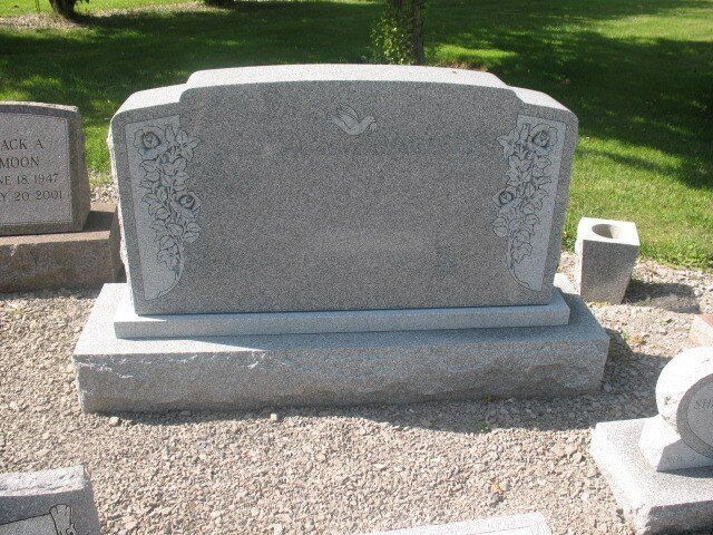 Gray granite gravestone with decorative carvings in a cemetery, set on gravel in sunlight