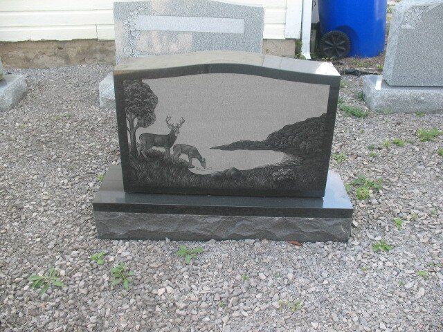 Black stone grave marker with engraved deer and tree silhouette on a gravel ground outdoors
