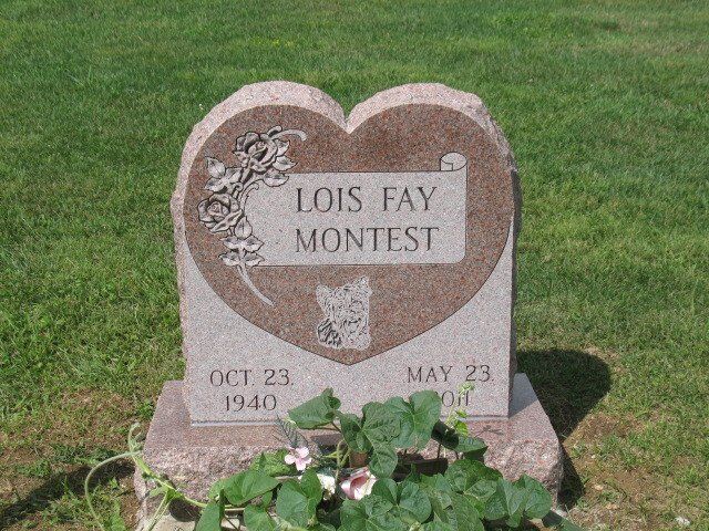 Heart-shaped gravestone in grass with pink flowers, engraved “LOIS FAY MONTEST” and dates beneath.