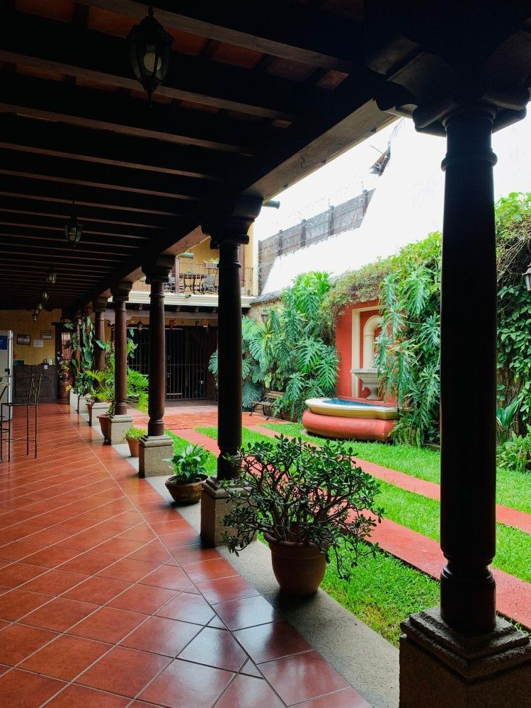 Un pasillo cubierto con baldosas de terracota, plantas en macetas, columnas oscuras y un jardín interior verde al fondo.