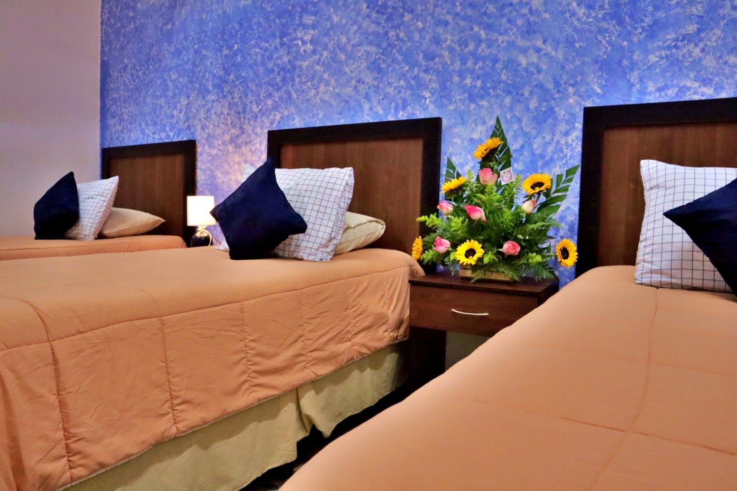 Dos camas individuales bien hechas con almohadas en una habitación, separadas por una mesita de noche con un arreglo floral.
