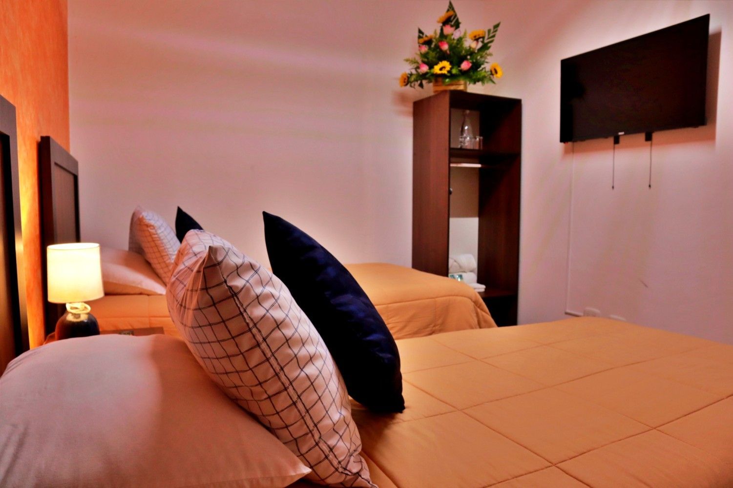 Habitación de hotel con dos camas bien hechas, iluminación cálida, televisor montado en la pared y un jarrón de flores en una repisa.