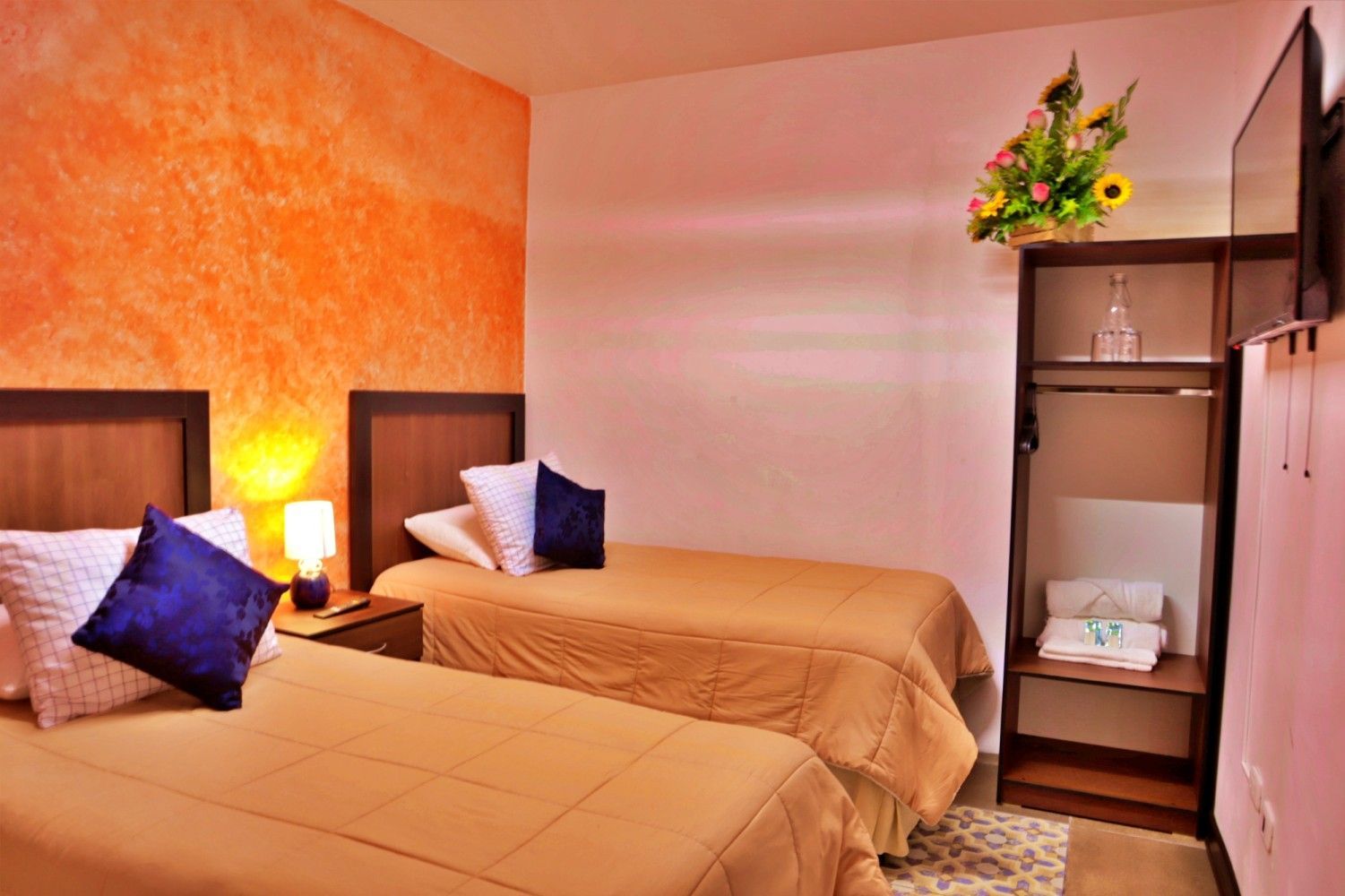 Dos camas individuales en una acogedora habitación de hotel con pared decorada en color naranja, lámpara de noche y armario abierto.