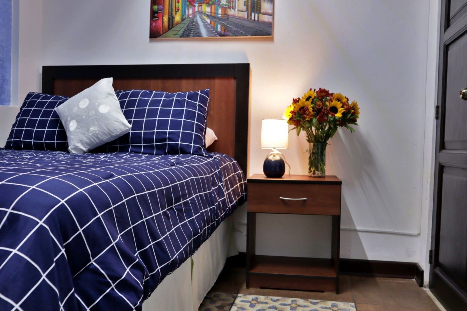 Dormitorio con colcha de cuadros azul marino, lámpara de noche, flores y cuadro enmarcado.