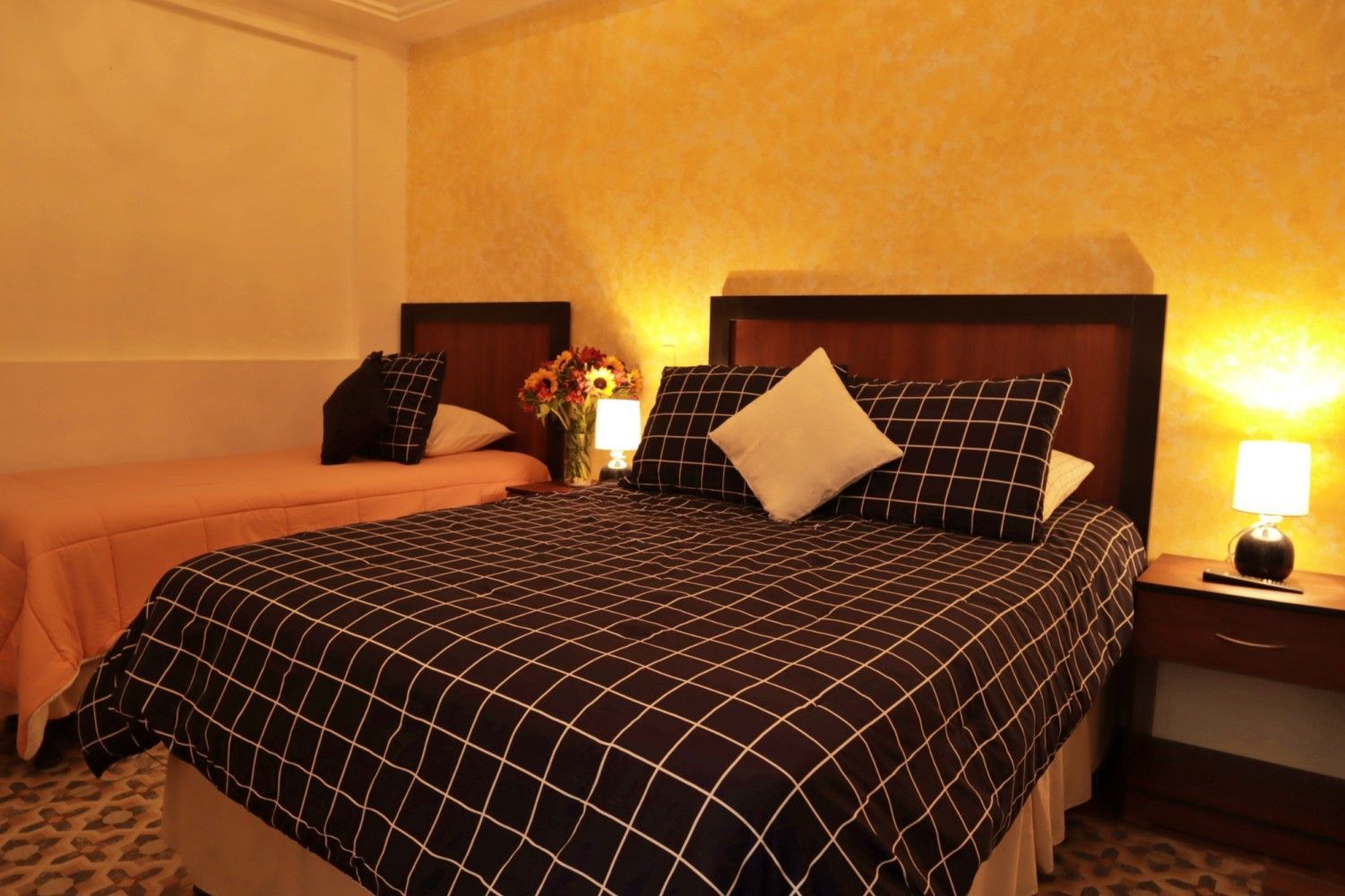 Habitación de hotel con dos camas, colchas a cuadros negras y lámparas de noche cálidas.