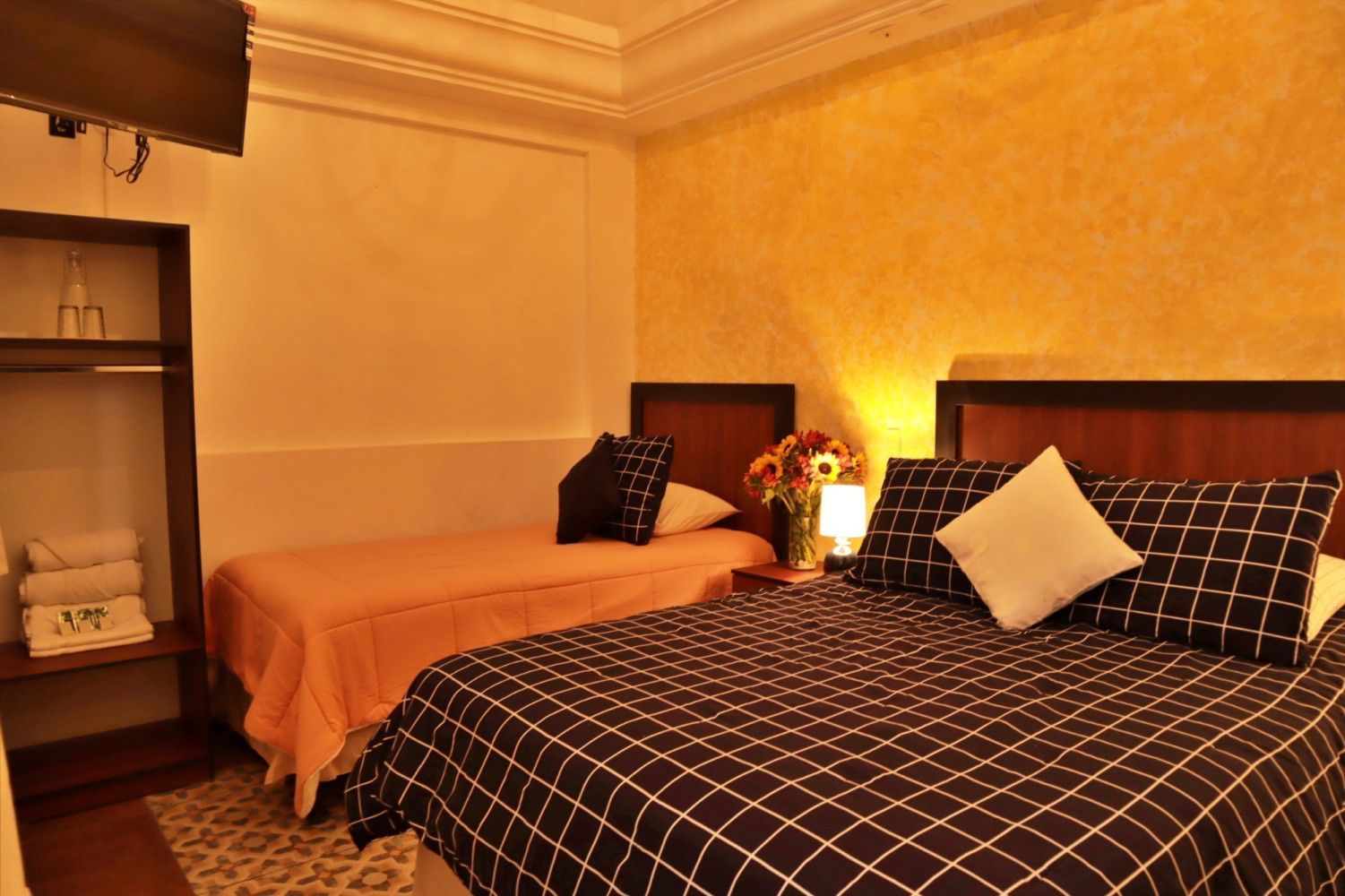 Acogedora habitación de hotel con dos camas, paredes de color beige cálido, ropa de cama estampada y un jarrón con girasoles.
