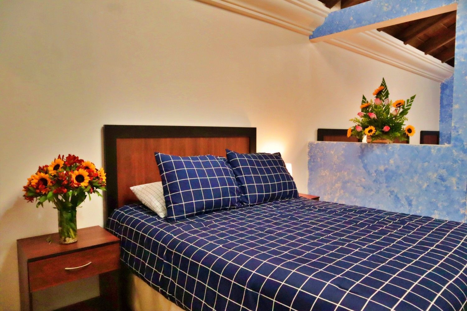 Acogedora habitación con colcha de cuadros azules, mesita de noche de madera, flores y techo inclinado con forma de manos.