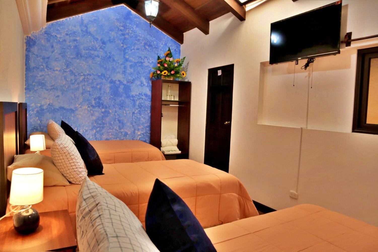 Acogedora habitación con dos camas, pared decorativa azul, vigas de madera, televisor montado en la pared e iluminación cálida con lámparas.